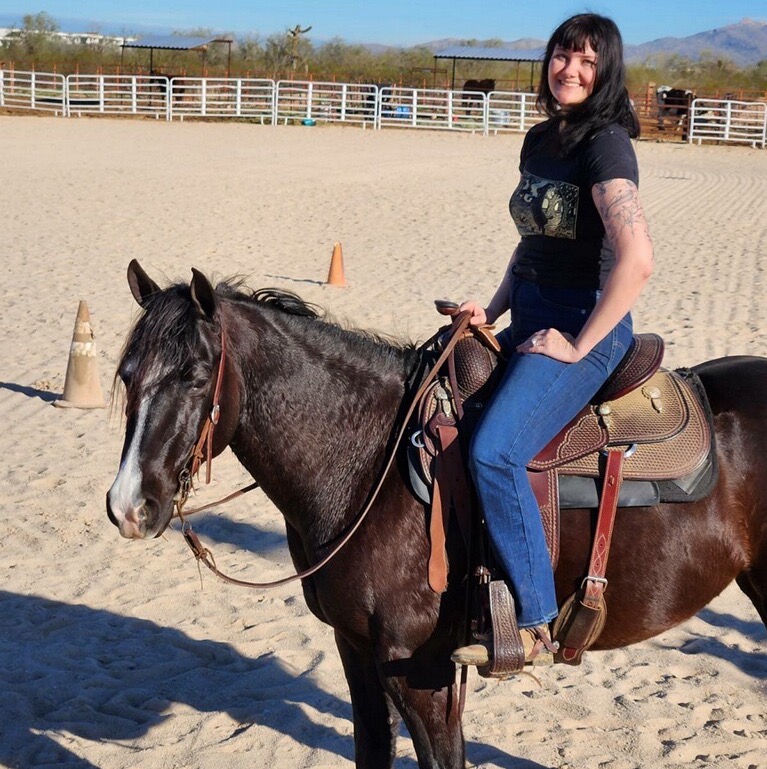 Image of a woman with dark hair sitting on a horse and smiling at the camera 